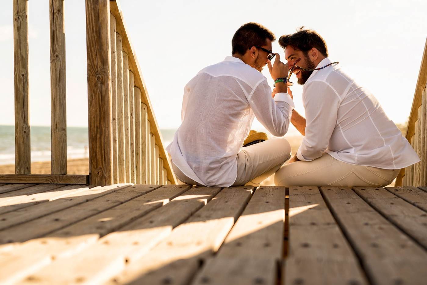 Two men in white shirts sitting on a wooden beach boardwalk laughing together at golden hour