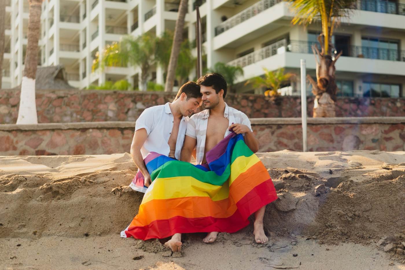 Two men sitting close on a sandy beach with a rainbow Pride flag across their laps