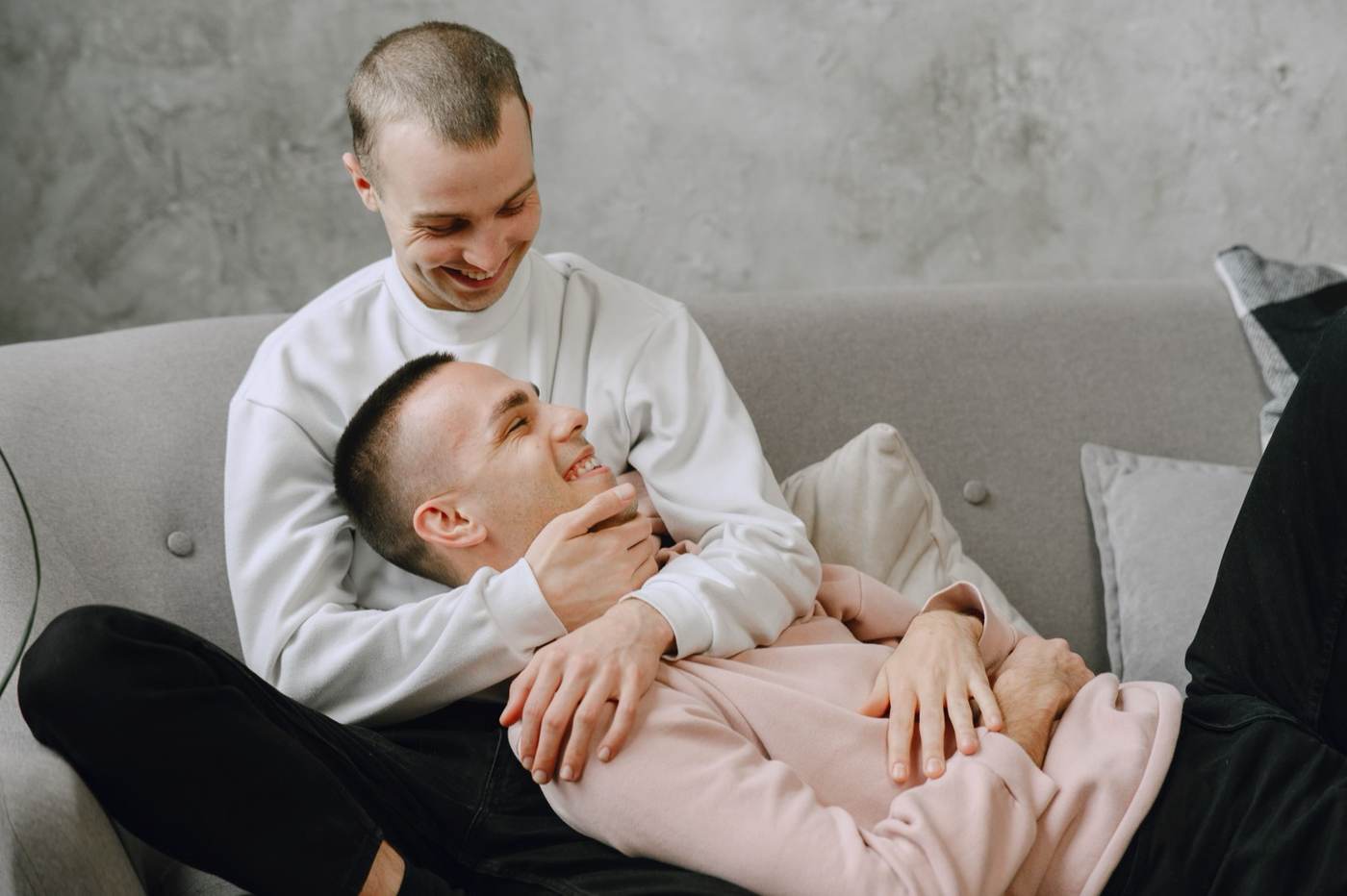 Two men cuddling on a grey sofa, one resting his head in the other's lap with a warm smile
