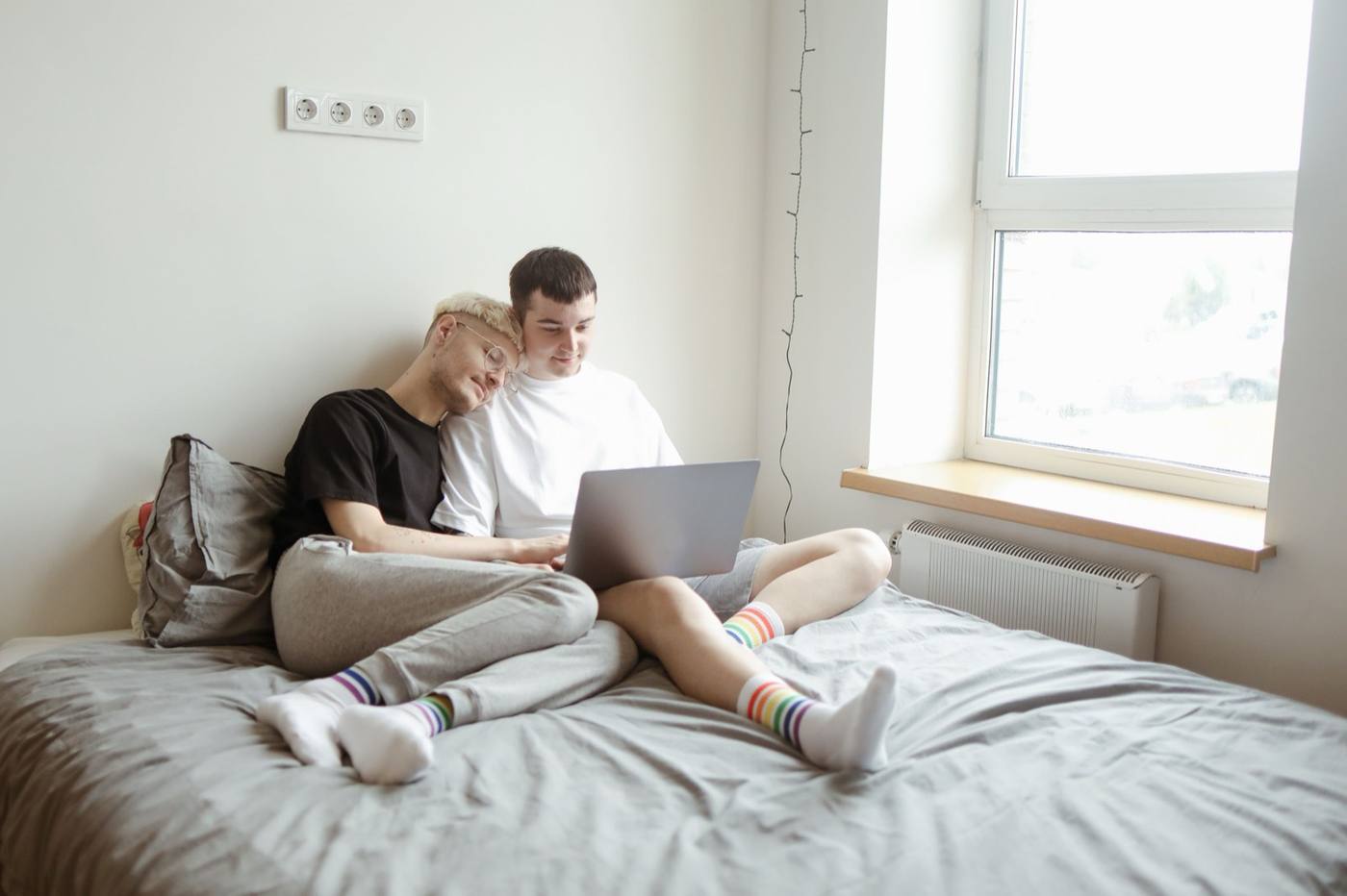 Two men sitting together on a bed looking at a laptop, wearing rainbow-stripe socks