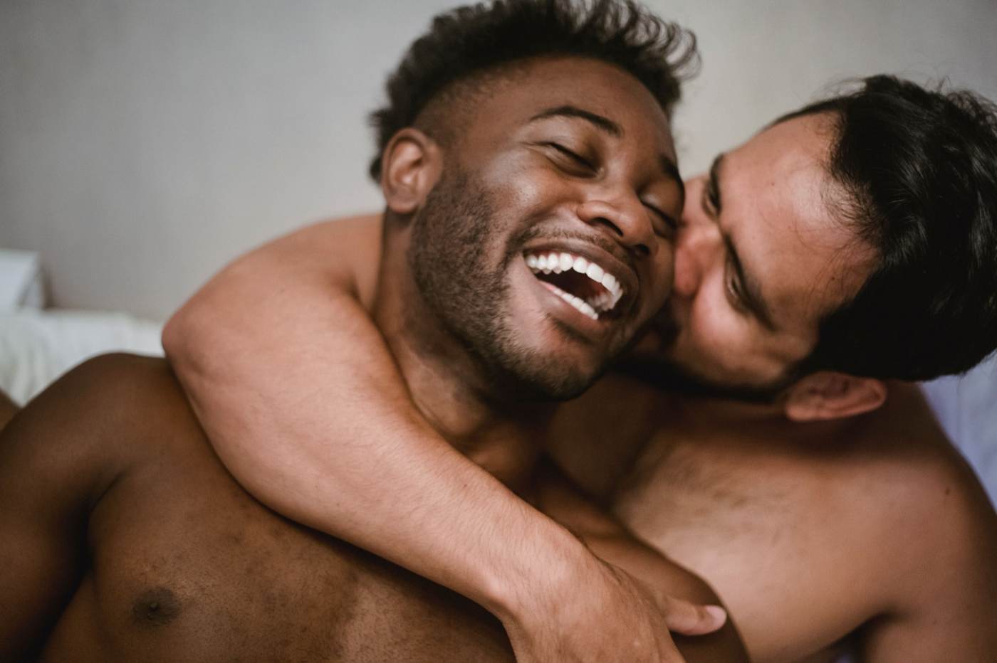Two shirtless men sharing a joyful cheek kiss while laughing