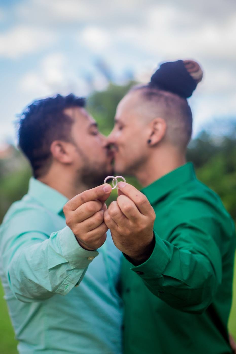 Two gold wedding bands held in focus with two men kissing softly in the background outdoors