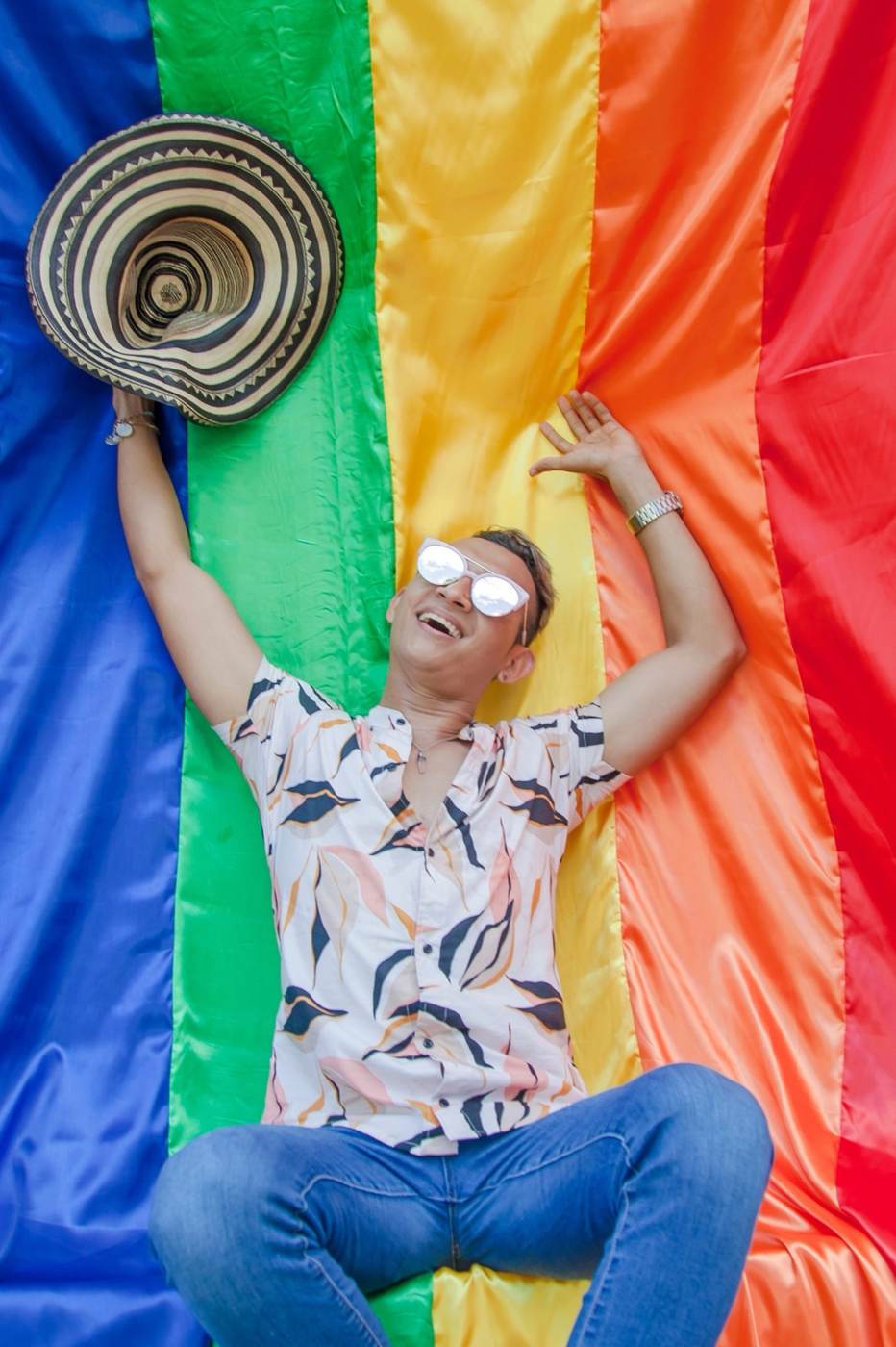 A joyful man sitting on a large rainbow Pride flag outdoors and smiling toward the camera